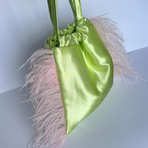 Op de afbeelding: Een limoengroene satijnen trekkoordtas met lichtroze veren. De tas heeft bijpassende groene satijnen handvatten. De tas is een klein, stijlvol accessoire.