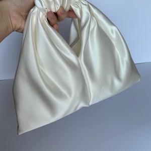 May include: Ivory satin handbag with a knotted handle. The bag has a soft, draped design and a smooth, lustrous finish. The bag is held by a hand, showcasing its shape and texture. The bag is a neutral color, suitable for various outfits.