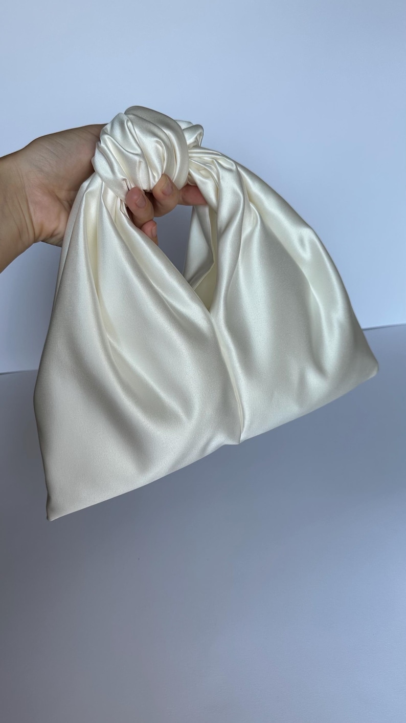 May include: Ivory satin handbag with a knotted handle. The bag has a soft, draped design and a smooth, lustrous finish. The bag is held by a hand, showcasing its shape and texture. The bag is a neutral color, suitable for various outfits.