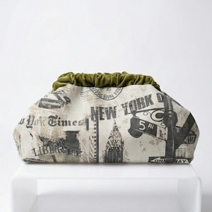 Peut inclure: Pochette avec un imprimé journal de New York. Le tissu blanc cassé présente du texte et des images en noir et gris de monuments. Le bord supérieur est en velours vert olive froncé.