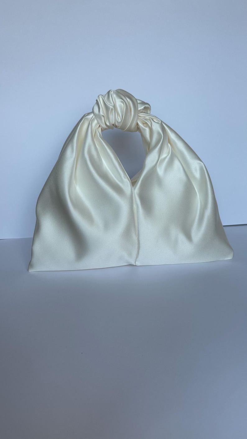 May include: Ivory satin handbag with a knotted handle. The bag has a trapezoid shape with a rounded top and a smooth, lustrous finish. The handle is formed by a knot, creating a circular opening. The bag is set against a white background.