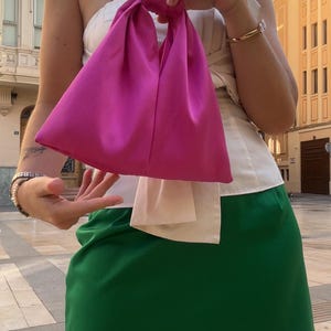 May include: A bright pink satin handbag with a knotted handle. The bag is held in front of a person wearing a white top and a green skirt. The background shows a city street with buildings.