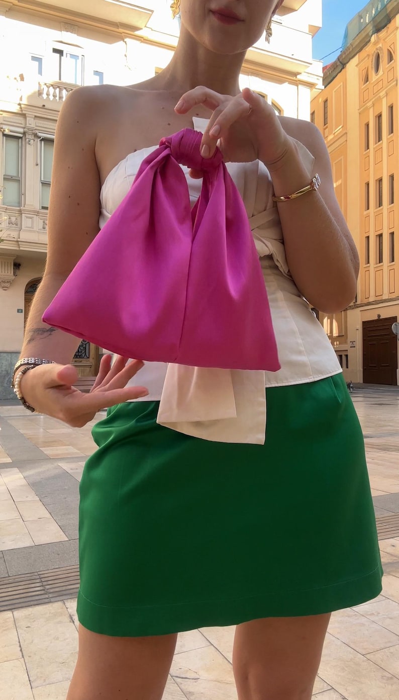 May include: A bright pink satin handbag with a knotted handle. The bag is held in front of a person wearing a white top and a green skirt. The background shows a city street with buildings.