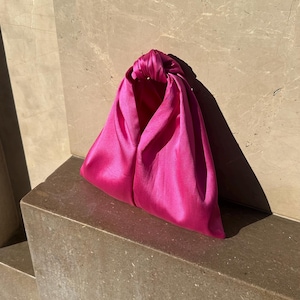 May include: A vibrant fuchsia satin clutch bag. The bag is triangular in shape, with a knotted handle at the top. The smooth, shiny fabric catches the light, creating highlights and shadows. The bag sits on a stone ledge.