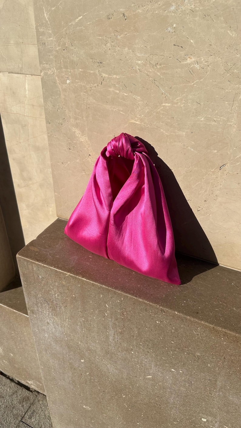 May include: A vibrant fuchsia satin clutch bag. The bag is triangular in shape, with a knotted handle at the top. The smooth, shiny fabric catches the light, creating highlights and shadows. The bag sits on a stone ledge.