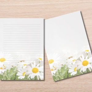 May include: Two sheets of white stationery with a daisy floral design. One sheet is lined for writing, and the other is blank. The daisy design features white petals and yellow centres with green leaves.