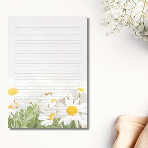 May include: A lined notepad with a floral design featuring white daisies with yellow centres and green leaves at the bottom. The notepad is on a white surface, with a cup of coffee and flowers in the background.