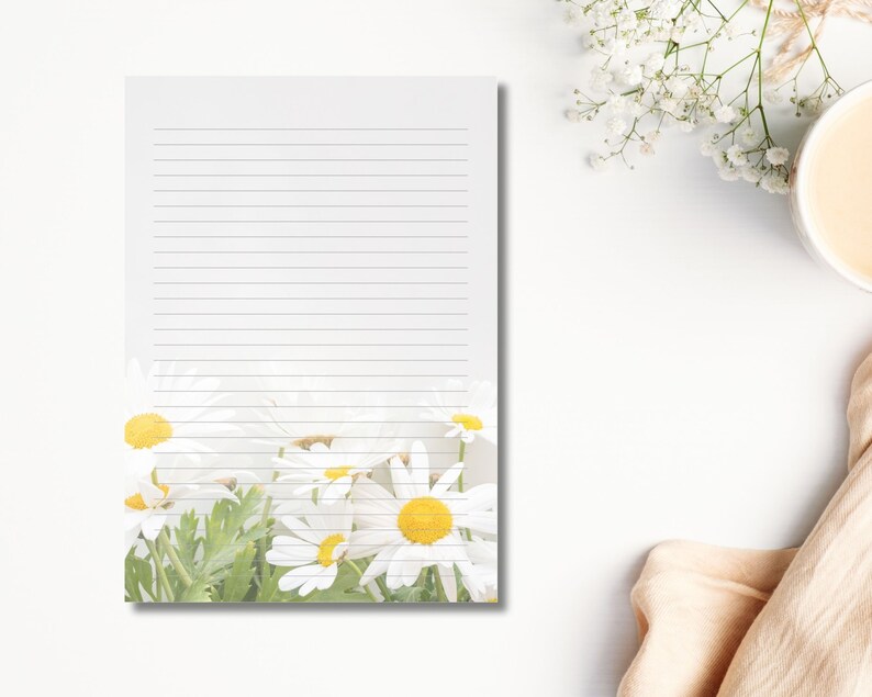 May include: A lined notepad with a floral design featuring white daisies with yellow centres and green leaves at the bottom. The notepad is on a white surface, with a cup of coffee and flowers in the background.