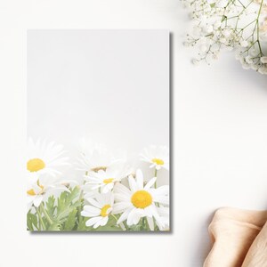 May include: A white notepad with a floral design featuring daisies with yellow centres and white petals. The notepad is surrounded by white flowers, a cup of coffee, and a beige cloth on a white surface.