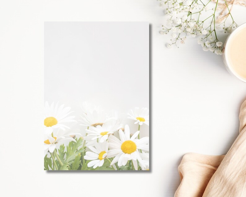 May include: A white notepad with a floral design featuring daisies with yellow centres and white petals. The notepad is surrounded by white flowers, a cup of coffee, and a beige cloth on a white surface.