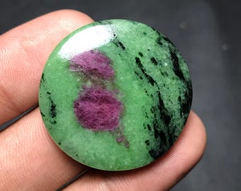May include: A round gemstone with a polished surface. The stone features a vibrant green base color with dark black veining and two prominent patches of deep pink-red. The gemstone is held in a hand.