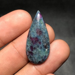 Puede incluir: Una gema en forma de lágrima con una superficie pulida. La piedra presenta una mezcla de tonos turquesa, morado intenso y verde. La gema se sostiene entre los dedos, mostrando su belleza natural y sus variaciones de color únicas.