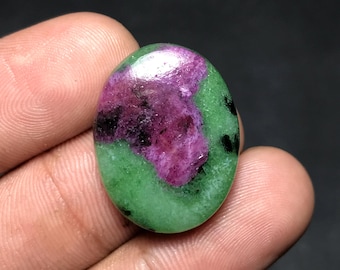 May include: Oval-shaped gemstone featuring a vibrant mix of colors. The stone showcases a central area of deep magenta, surrounded by a bright green border. Small black inclusions are scattered throughout the green.