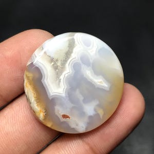 Puede incluir: Una gema redonda y pulida con un patrón arremolinado de tonos blancos, grises y amarillos. La piedra tiene una superficie lisa y un aspecto natural. La gema se sostiene en una mano.