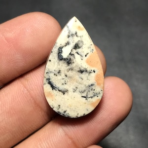 Op de afbeelding: Een druppelvormige edelsteen met een crèmekleurige basis, bespikkeld met zwart en geaccentueerd met perzikkleurige vlekken. De gepolijste steen wordt in een hand gehouden en toont zijn unieke patroon en natuurlijke schoonheid.