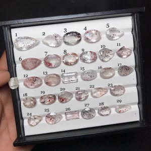 以下が含まれることがあります： 赤と黒の鉱物インクルージョンを含む、29個のファセットカットされた透明な宝石が入った黒いディスプレイボックス。各石には1から29までの番号が付けられ、白い溝付きのインサートの上に列状に配置されています。