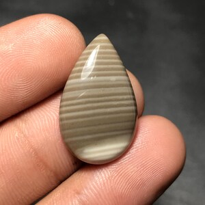 May include: A polished, teardrop-shaped gemstone. It displays a striped pattern of light brown and off-white bands. The stone is held between fingers. The gemstone's surface reflects light.