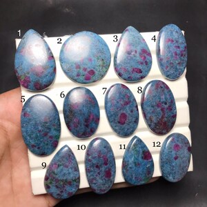 Op de afbeelding: Twaalf gepolijste cabochon edelstenen gerangschikt op een wit displaybord, genummerd van één tot twaalf. De stenen hebben een gevlekte blauwe basiskleur met ingebedde magenta en donkerroze insluitingen, in diverse ovale en druppelvormen.