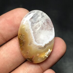 Puede incluir: Gema ovalada con superficie pulida. La piedra presenta una mezcla de colores blanco, beige y marrón, con una zona translúcida en la parte superior. La gema se sostiene entre los dedos, mostrando sus patrones y texturas naturales. Ideal para joyería.