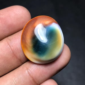 May include: A polished, oval gemstone with a vibrant color gradient. The stone transitions from a warm orange-brown at the edges to a mix of white, blue, and green in the center. The gemstone is held in a hand.