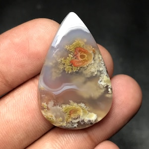 May include: A polished, teardrop-shaped gemstone. The stone displays a mix of white, grey, and brown hues, with a central pattern of orange and yellow. The design resembles a floral or cloud-like formation. The surface is smooth, reflecting light.