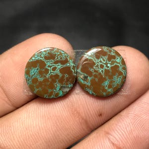 Op de afbeelding: Twee ronde, gepolijste edelstenen met een bruin en turquoise patroon. De stenen worden bij elkaar gehouden met doorzichtige tape. De edelstenen hebben een unieke, natuurlijke uitstraling, met een netwerk van turquoise aderen tegen een bruine achtergrond.