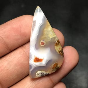 May include: A polished, triangular gemstone with a mix of colors. The stone features white, gray, orange, and brown patterns. The gemstone has a smooth, glossy surface, and is likely used for jewelry or collection.