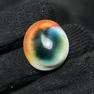 Pode incluir: Uma pedra preciosa oval polida com um padrão de cores único. A pedra apresenta uma mistura de cores, incluindo laranja, azul, verde e branco, criando um efeito de redemoinho, semelhante a um olho. A pedra preciosa está colocada sobre um fundo escuro.