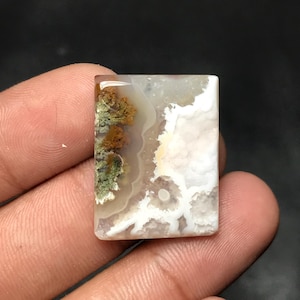 Puede incluir: Una gema rectangular con un patrón único. La piedra presenta una mezcla de colores, incluyendo blanco, marrón y verde, con una apariencia translúcida. El diseño se asemeja a un paisaje o arte abstracto, lo que la convierte en una pieza distintiva.