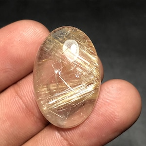May include: Oval-shaped, translucent gemstone with golden, needle-like inclusions. The stone has a polished surface and a light brown hue, with a bright, reflective quality. The inclusions create a striking visual effect, resembling strands of gold.