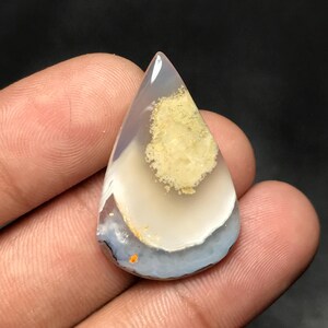 Puede incluir: Una gema pulida en forma de lágrima. La piedra presenta capas de blanco, azul claro y gris, con un parche central de color amarillo verdoso. La gema se sostiene en una mano.