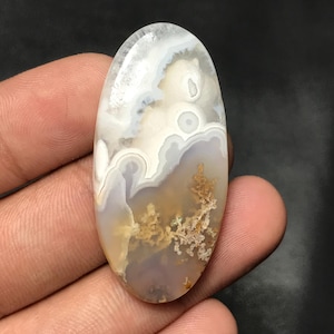May include: An oval gemstone with a distinctive pattern of white, grey, and brown swirls. The polished stone has a natural, organic appearance. The colours blend, creating a marbled effect. The gemstone is held in a hand.