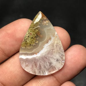 Pode incluir: Uma pedra preciosa em forma de gota com uma mistura de cores. A parte superior apresenta inclusões verdes e castanhas semelhantes a musgo, que transitam para áreas brancas e translúcidas. A pedra tem uma superfície polida.