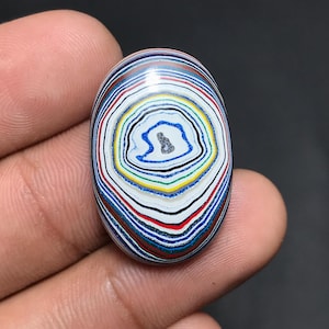 May include: An oval-shaped stone with a unique pattern of concentric, colorful rings. The design features layers of red, white, blue, yellow, and black, creating a striking visual effect. The stone is smooth and polished, with a glossy finish.