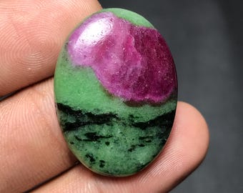 May include: Oval-shaped gemstone with a vibrant color palette. The top section features a deep magenta hue, transitioning to a bright green in the middle, and a band of black at the bottom. The stone is held in a hand.