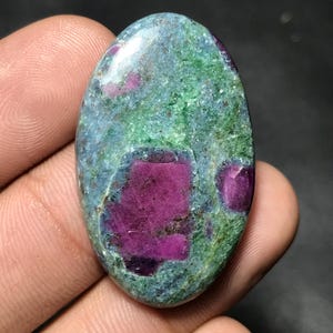 May include: An oval-shaped gemstone with a mix of colors. The stone features a base of teal and green hues, with prominent patches of deep magenta. The polished surface of the stone reflects light, highlighting its natural patterns and textures.