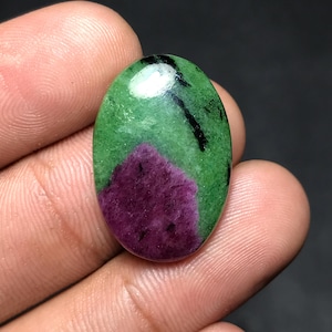 Puede incluir: Gema de forma ovalada con una vibrante combinación de colores. La piedra presenta una base de color magenta intenso, que se transforma en un verde brillante con vetas negras. La superficie pulida refleja la luz, resaltando la belleza natural.