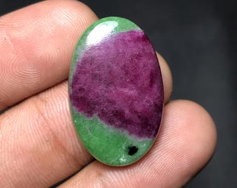 May include: An oval-shaped gemstone with a vibrant color combination. The stone features a deep magenta center, transitioning to a bright green border. A small black inclusion is visible near the edge. The polished surface reflects light, highlighting the stone's natural beauty.