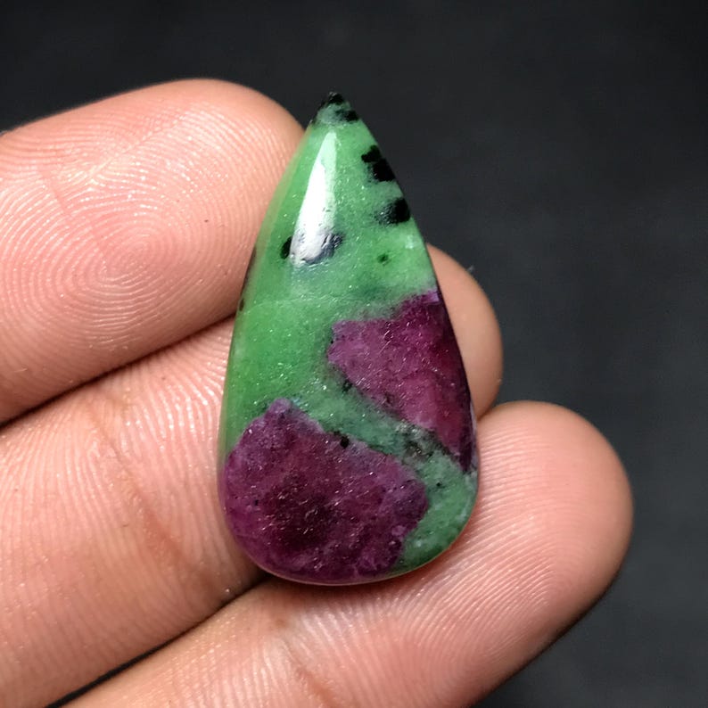 May include: A teardrop-shaped gemstone featuring a vibrant mix of colors. The stone showcases a base of vivid green, interspersed with patches of deep magenta and flecks of black. The polished surface reflects light, highlighting the natural patterns.