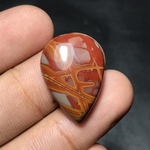 Puede incluir: Una gema pulida en forma de l&aacute;grima con un llamativo patr&oacute;n de colores rojo, naranja y crema. La piedra tiene un acabado brillante y se sostiene en una mano, mostrando su belleza natural y veteado &uacute;nico.