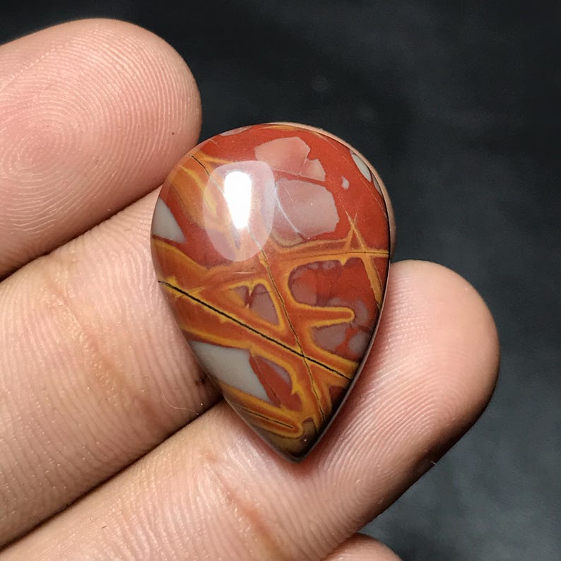Puede incluir: Una gema pulida en forma de l&aacute;grima con un llamativo patr&oacute;n de colores rojo, naranja y crema. La piedra tiene un acabado brillante y se sostiene en una mano, mostrando su belleza natural y veteado &uacute;nico.