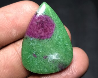 May include: A teardrop-shaped gemstone featuring a vibrant combination of colors. The stone showcases a deep magenta-colored circular inclusion set within a textured green base. The polished surface reflects light, highlighting the natural patterns.