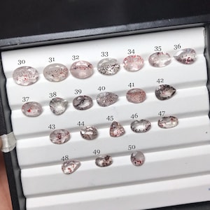 Puede incluir: Una colección de veintiuna piedras preciosas facetadas dispuestas en una bandeja blanca. Cada piedra es transparente con inclusiones minerales rojas, marrones o negras. Las piedras están numeradas del 30 al 50 y presentan formas ovaladas y de pera.