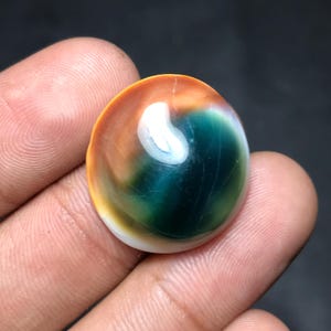 Pode incluir: Uma pedra preciosa redonda e polida com um padrão de cores em espiral. A gema apresenta uma mistura de tons laranja, branco e verde escuro. A pedra é segurada em uma mão, mostrando sua superfície lisa e reflexiva. A pedra preciosa é um item natural único.