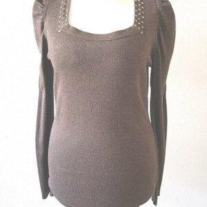 Könnte beinhalten: Ein brauner Langarmpullover mit quadratischem Ausschnitt. Der Ausschnitt und die Schulternähte sind mit kleinen, silberfarbenen Nieten verziert. Der Pullover hat eine taillierte Silhouette und Puffärmel, was einen klassischen und eleganten Look erzeugt.