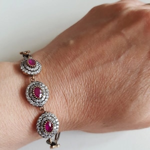 Könnte beinhalten: Goldfarbenes Armband mit drei ovalen, rubinroten Edelsteinen, die jeweils von kleinen, klaren Kristallen umgeben sind. Das Armband ist durch filigrane Glieder verbunden und wird am Handgelenk getragen.