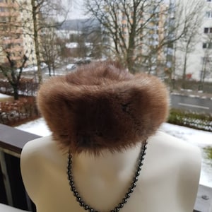 Könnte beinhalten: Ein brauner Pelz Hut mit breiter Krempe. Der Hut befindet sich auf einem weißen Mannequin mit einer schwarzen Perlenkette.