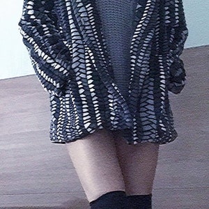 May include: A black and white patterned coat with a textured design. The coat is worn over a gray sweater. The person is also wearing black knee-high socks.