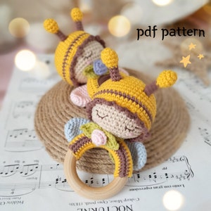 May include: Two crocheted bee-themed baby rattles with wooden rings. The bees are yellow and brown with embroidered eyes and a pink flower. The text "pdf pattern" is visible in the image.