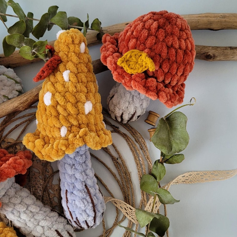 May include: Three crocheted mushroom toys with different coloured tops. One is red, one is yellow with white dots, and one is blue. The mushrooms are sitting on a background of twine, lace, and greenery.